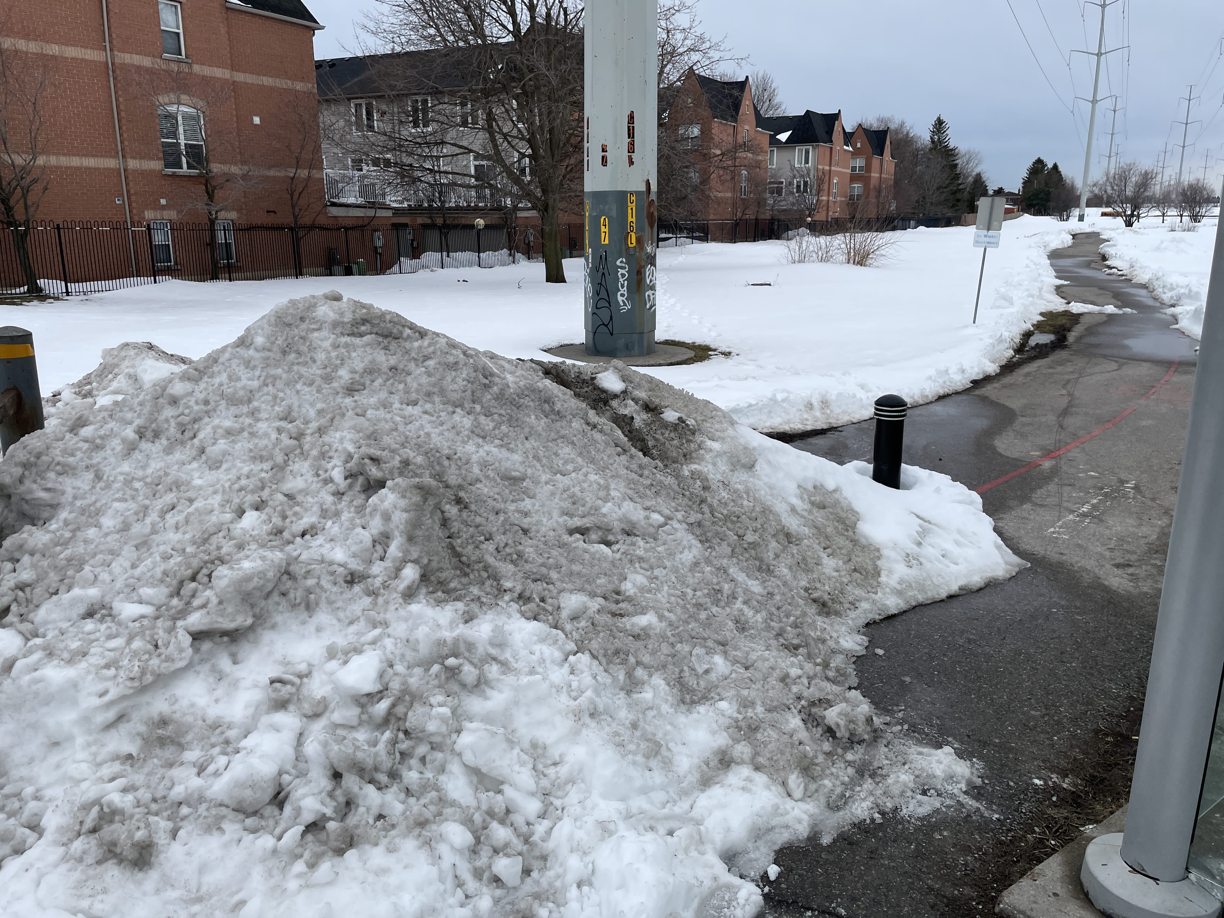 Photo of a trail entrance from Lawrence Ave E, with a pile of dirty snow about 1.5 metres tall and 4 metres wide. The passable gap between the pile of snow and a bus shelter is about half a metre wide. Power lines and houses are visible in the background.